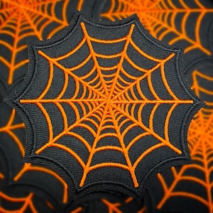May include: A black embroidered spiderweb patch with orange thread. The patch is octagonal and features a detailed web design. The background shows a pattern of similar spiderwebs, creating a Halloween theme.