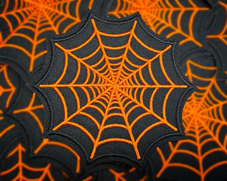 May include: A black embroidered spiderweb patch with orange thread. The patch is octagonal and features a detailed web design. The background shows a pattern of similar spiderwebs, creating a Halloween theme.