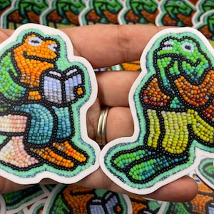 May include: Two colourful beaded frog stickers, one reading a book and the other with its hands on its chin. The frogs are green, orange, and blue.