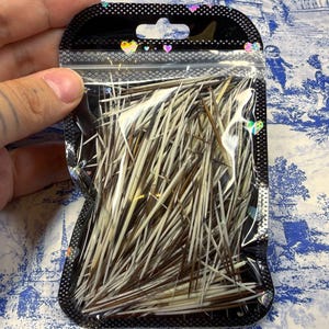 May include: A clear plastic bag filled with a collection of porcupine quills. The quills are a mix of brown and white, and are contained within a black plastic bag with a small hole at the top.