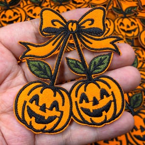 May include: Embroidered orange pumpkin patches with black outlines and smiling faces, connected by stems and a black bow. Green leaves accent the design, perfect for Halloween or fall-themed crafts.