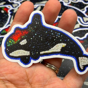 May include: A black and white orca whale sticker with red, green, and blue accents. The orca is made of small beads and is surrounded by a blue border.