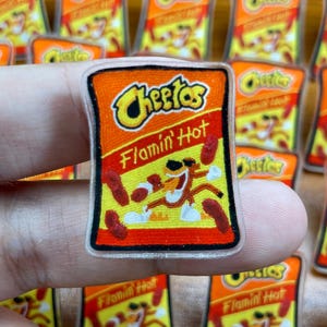 May include: A small, rectangular pin with the Cheetos Flamin' Hot logo and mascot, in orange and yellow. The pin is held in a hand, with other similar pins visible in the background.