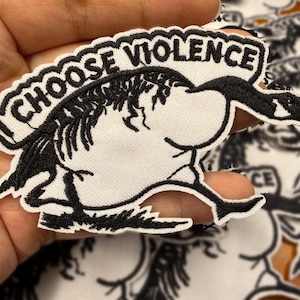 Może przedstawiać: Haftowana naszywka z postacią z kreskówki gęsi i napisem "CHOOSE VIOLENCE". Projekt jest czarno-biały, z gęsią w pozycji do biegu. Naszywka jest biała z czarnymi szwami. Prawdopodobnie do odzieży lub akcesoriów.