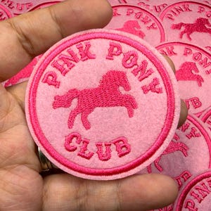 Puede incluir: Parche bordado redondo rosa con un caballo rosa y las palabras "PINK PONY CLUB". El parche tiene un borde y letras de color rosa más oscuro. Varios parches son visibles.