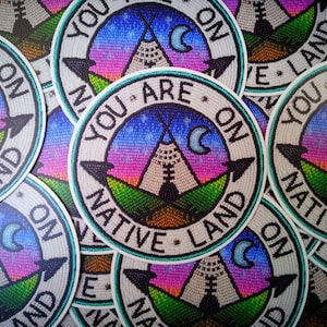 May include: A close-up of several round stickers with a beaded design. The stickers feature a teepee, a crescent moon, and the text "You Are On Native Land".