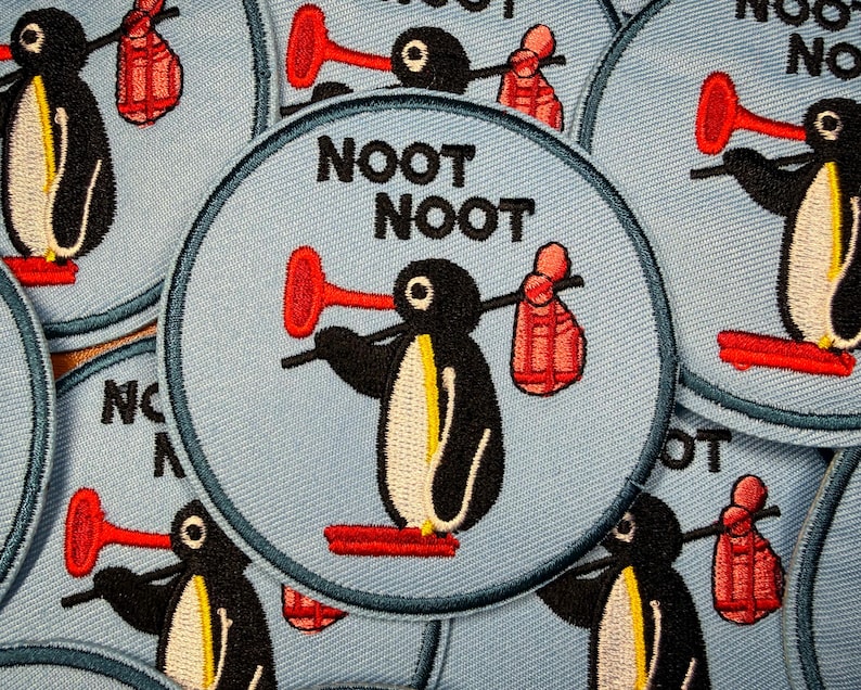 K&ouml;nnte beinhalten: Gestickter Aufn&auml;her mit einem Cartoon-Pinguin-Design. Der Pinguin hat einen roten P&ouml;mpel als Nase und tr&auml;gt eine rote Tasche. Der Text "NOOT NOOT" steht &uuml;ber dem Pinguin. Der Aufn&auml;her befindet sich auf hellblauem Hintergrund.