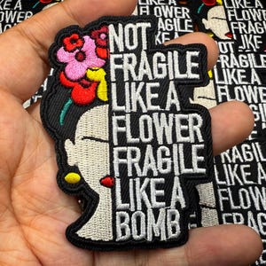 May include: Black embroidered patch with a portrait of a woman with floral headwear. The patch reads "NOT FRAGILE LIKE A FLOWER FRAGILE LIKE A BOMB" in white text.