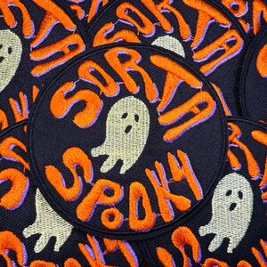 以下が含まれることがあります： 「SORTA SPOOKY」と書かれたオレンジ色の刺繍文字とゴーストグラフィックが特徴の黒い円形パッチ。テキストは紫色の輪郭です。ゴーストは白です。パッチは積み重ねられています。