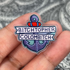 May include: A clear acrylic pin featuring a stylized anchor design with the text "BITCHTOPHER COLOMBITCH" in a banner. The anchor is purple, teal, and blue with red accents.