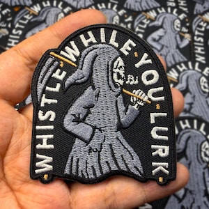 May include: Black patch featuring a gray Grim Reaper playing a flute, with the text "WHISTLE WHILE YOU LURK". The patch has a black border with gold detailing, suitable for clothing or bags.