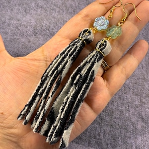 Pode incluir: Um par de brincos compridos com borlas. Os brincos apresentam ganchos dourados, contas em forma de flor azul claro e verde, e borlas listradas em preto e branco. As borlas são feitas de tecido e são presas com arame dourado.