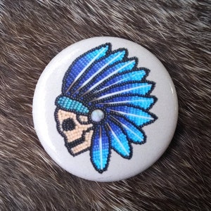 May include: A white round button with a beaded design of a Native American headdress in blue and white colors. The headdress is worn by a skull with a beaded design.
