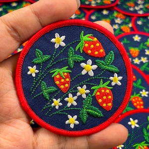 Può includere: Toppa ricamata con un design a fragola. La toppa ha uno sfondo blu navy e un bordo rosso. Il design include fragole rosse con semi gialli, fiori bianchi e foglie verdi.