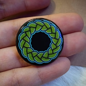 Puede incluir: Un botón redondo con fondo negro y un diseño tejido verde y azul.