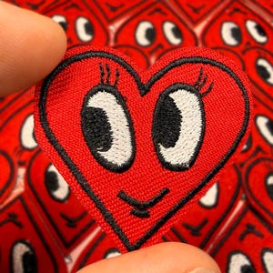 May include: A red heart-shaped embroidered patch with a smiling face, black outline, and white eyes. The patch is held between two fingers, with a background of similar patches. The patch has a cute, cartoonish design.