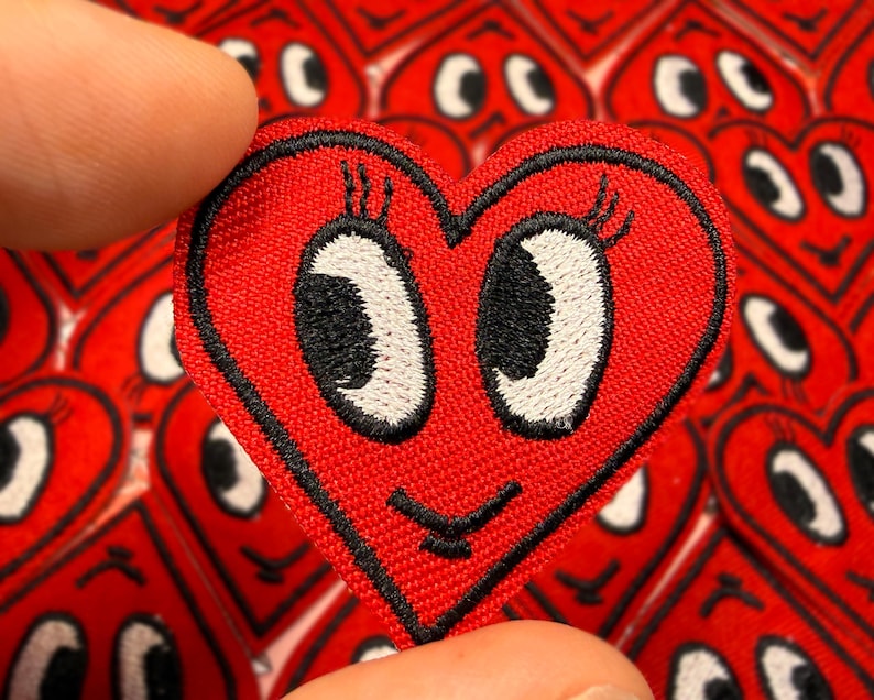 May include: A red heart-shaped patch with a smiling face, black outline, and white eyes. The patch is held between two fingers, with a background of similar patches. The patch has a cute, cartoonish design.