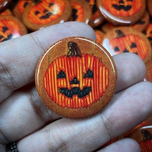 May include: A close-up of a round, wooden button featuring a stitched image of a jack-o'-lantern. The jack-o'-lantern is orange with a black stitched face.