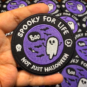 May include: Circular black and purple patch with the text "Spooky For Life, Not Just Halloween." The patch features a white ghost, bats, a skull, and the word "Boo" in a speech bubble. The patch is held in a hand.