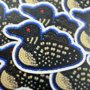 May include: A sticker with a beaded design of a loon bird. The loon has a black body with white and tan accents, a red eye, and a blue outline. The sticker is on a white background.