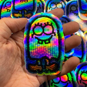 May include: Holographic sticker featuring a cartoon character with a smiling face, large eyes, and a toothy grin. The sticker has a rainbow color scheme and a beaded appearance. Additional stickers are visible in the background.