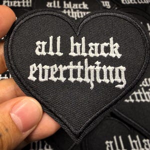 Könnte beinhalten: Ein schwarzer herzförmiger Aufnäher mit dem Schriftzug "all black everything" in weißen gotischen Buchstaben gestickt. Der Aufnäher wird vor einem Hintergrund ähnlicher Aufnäher gehalten.