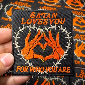 May include: Black square patch with orange text and imagery. The text reads "SATAN LOVES YOU" and "FOR WHO YOU ARE". The image features two orange hands forming a heart shape, surrounded by a barbed wire circle.