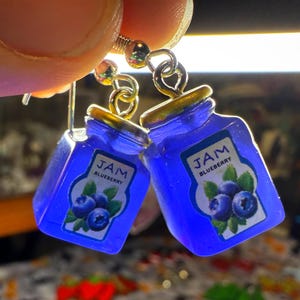 Könnte beinhalten: Ein Paar Ohrhänger mit kleinen blauen Marmeladengläsern. Jedes Glas hat einen goldfarbenen Deckel und ein Etikett mit der Aufschrift "JAM BLUEBERRY" und einer Blaubeer-Grafik. Die Ohrringe hängen an silberfarbenen Haken.