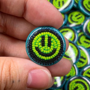 May include: A close-up of a teal blue and black beaded smiley face pin. The smiley face is outlined in black beads and has a green face with black eyes.