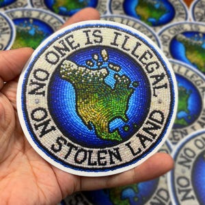 Puede incluir: Una pegatina redonda con un diseño de cuentas de América del Norte en colores azul, verde y blanco. El texto "No One Is Illegal On Stolen Land" está escrito en un círculo alrededor del mapa.