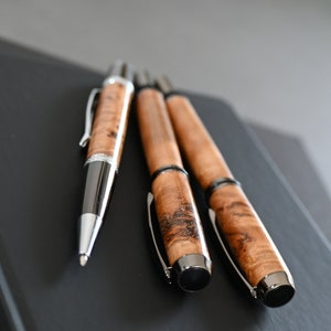 以下が含まれることがあります： 木製のバレルと銀のアクセントが付いた3本のペン。ペンは黒い表面に置かれています。