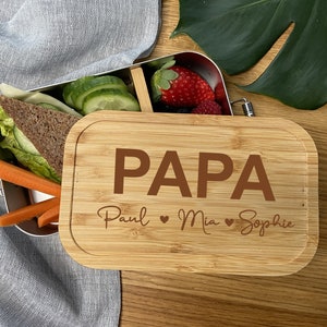 Könnte beinhalten: Eine rechteckige Edelstahl-Lunchbox mit einem Bambusdeckel. Der Deckel ist mit dem Wort PAPA in großen Buchstaben graviert, gefolgt von den Namen Paul, Mia und Sophie, die durch Herzsymbole getrennt sind. Die Box enthält ein Sandwich und Obst.