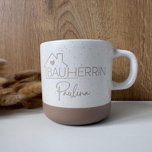 Könnte beinhalten: Eine gesprenkelte, weiße Keramik-Tasse mit braunem Boden und Henkel. Die Tasse trägt den Text "BAUHERRIN Paulina" in Hellgrau sowie eine Hausgrafik mit Herz.