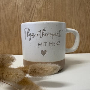 Könnte beinhalten: Ein weißer Keramikbecher mit gesprenkelter Oberfläche und einem beigefarbenen Boden. Auf der Vorderseite steht der graue Schriftzug Physiotherapeut MIT HERZ über einem kleinen grauen Herzsymbol. Der Becher hat einen geschwungenen Henkel.
