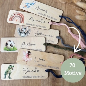 Könnte beinhalten: Holzlesezeichen mit farbenfrohen Illustrationen von Rakete, Regenbogen, Fledermaus, Fußball, Fee und Dinosaurier. Jedes Lesezeichen hat eine Quaste und den Text "SCHULKIND 2025 - DU BIST ERSTE KLASSE".