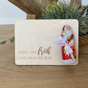 Könnte beinhalten: Ein Holzschild mit dem Text "Lasst uns froh und munter sein" und einer Schokoladen-Weihnachtsmann-Figur mit einem rosa Band.