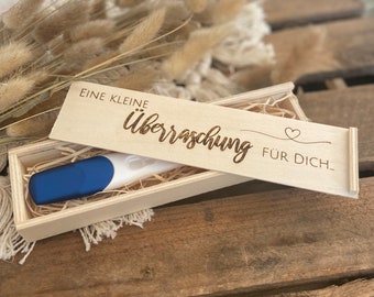May include: A wooden box with the text "Eine kleine Überraschung für dich..." engraved on the lid. The box contains a pregnancy test.