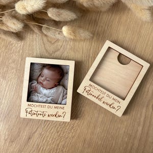 Könnte beinhalten: Zwei quadratische Holzfotorahmen. Einer enthält ein Babyfoto, der andere ist leer. Beide Rahmen tragen den deutschen Text "Möchtest du meine Patentante werden?" eingraviert. Die Rahmen liegen auf einer Holzoberfläche.
