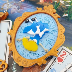 Peut inclure: Pendentif doré représentant Alice au pays des merveilles tombant dans un miroir. Alice porte une robe bleue et a les cheveux blonds. Le fond est bleu clair avec des illustrations et du texte blancs. Des cartes à jouer sont visibles en arrière-plan.