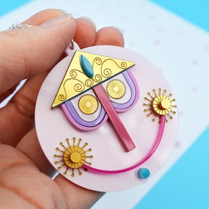 May include: A round pendant with a whimsical face design. The pendant features a pink background with a gold and teal triangle, purple and gold details, and a pink smile. Two gold sunbursts and a teal dot complete the design.