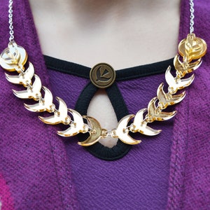 May include: A gold necklace with a crescent moon design. The necklace is made of metal and has a silver chain. The necklace is worn by a person with a purple shirt.