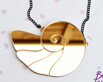 May include: A gold-colored, shell-shaped pendant necklace with a black chain. The pendant has a spiral design and is made of a shiny, reflective material. The necklace is displayed on a light background with pink accents. The logo "Bobbins Inspired" is visible.