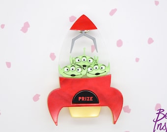 May include: A red and yellow rocket ship with a clear plastic top. Inside the rocket are three green alien figures with three eyes each. The rocket has the word "PRIZE" on the front.