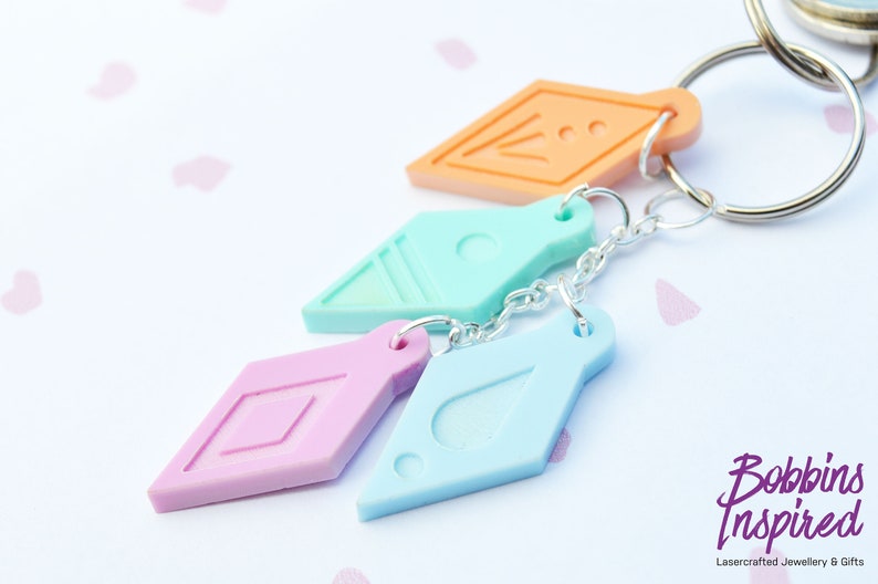 Peut inclure: Un ensemble de quatre porte-cl&eacute;s en forme de diamant de couleurs diff&eacute;rentes avec des motifs diff&eacute;rents. Les porte-cl&eacute;s sont attach&eacute;s &agrave; une cha&icirc;ne argent&eacute;e et un anneau de cl&eacute; argent&eacute;. Les porte-cl&eacute;s sont dans des tons de rose, bleu et orange.