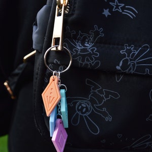 Peut inclure: Un tirette de fermeture &eacute;clair dor&eacute;e avec un porte-cl&eacute;s attach&eacute; &agrave; un sac noir avec un motif de dessin anim&eacute; blanc. Le porte-cl&eacute;s est orn&eacute; de plusieurs breloques color&eacute;es, dont un breloque en forme de diamant, un breloque carr&eacute; et un breloque rectangulaire.