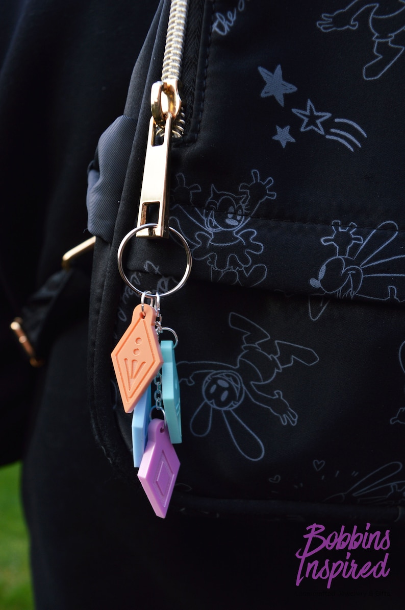 Peut inclure: Un tirette de fermeture &eacute;clair dor&eacute;e avec un porte-cl&eacute;s attach&eacute; &agrave; un sac noir avec un motif de dessin anim&eacute; blanc. Le porte-cl&eacute;s est orn&eacute; de plusieurs breloques color&eacute;es, dont un breloque en forme de diamant, un breloque carr&eacute; et un breloque rectangulaire.