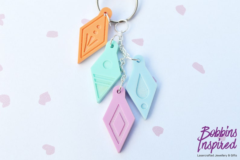 Peut inclure: Un porte-cl&eacute;s avec quatre breloques en forme de diamant de couleurs diff&eacute;rentes. Les breloques sont orange, bleu clair, bleu clair avec un motif de goutte blanche et rose. Le porte-cl&eacute;s est attach&eacute; &agrave; un anneau de cl&eacute; argent&eacute;.