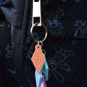 Peut inclure: Un tirette de fermeture &eacute;clair dor&eacute;e avec un porte-cl&eacute;s attach&eacute; &agrave; un ensemble de quatre breloques color&eacute;es. Les breloques sont en forme de diamants et sont dans des tons de rose, bleu et orange.