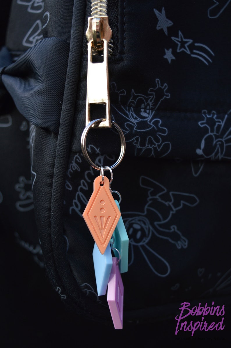 Peut inclure: Un tirette de fermeture &eacute;clair dor&eacute;e avec un porte-cl&eacute;s attach&eacute; &agrave; un ensemble de quatre breloques color&eacute;es. Les breloques sont en forme de diamants et sont dans des tons de rose, bleu et orange.