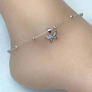 May include: A silver anklet featuring a butterfly charm with white and black accents. The delicate chain is adorned with small, round beads. This piece of jewelry is designed to be worn around the ankle.